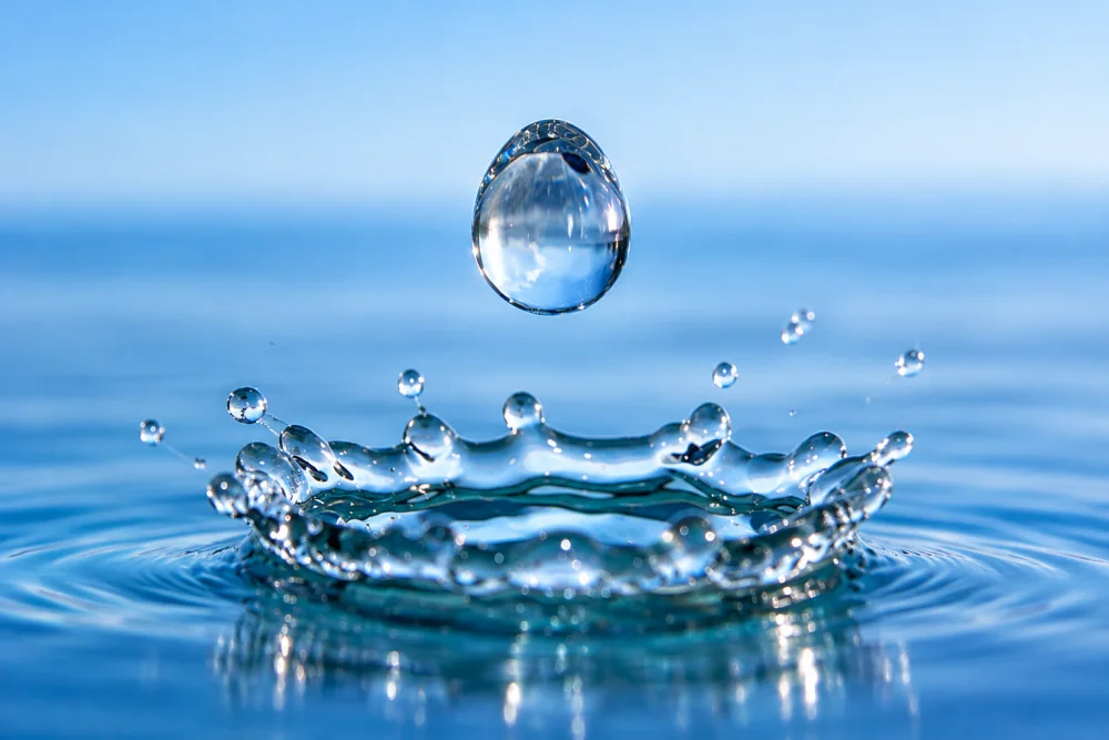 Goutte d'eau ronde et transparente en suspension au-dessus d'une onde circulaire, sur fond d'eau bleue avec de petites gouttelettes autour.